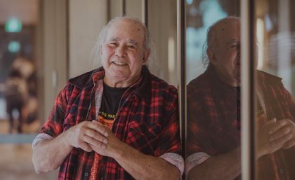 Kev Carmody is a 2022 UQ Honorary Doctorate recipient. a man in a check shirt leans against a window in which he is reflected